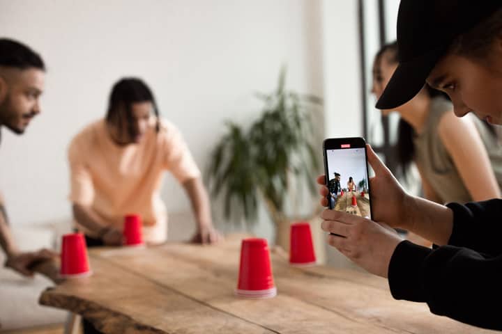 Group of young adults engaging in a creative game with red cups indoors, capturing moments on a smartphone.