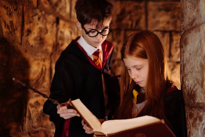 Two children in wizard costumes reading a spellbook in a dimly lit stone room.