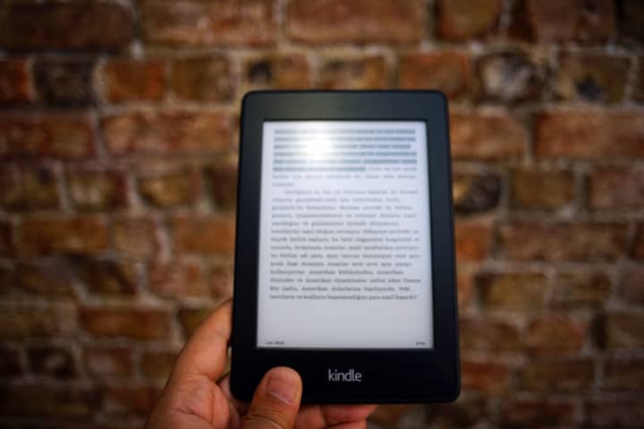 Close-up of a Kindle e-reader held by a person, showcasing a digital reading device against a textured brick wall backdrop.