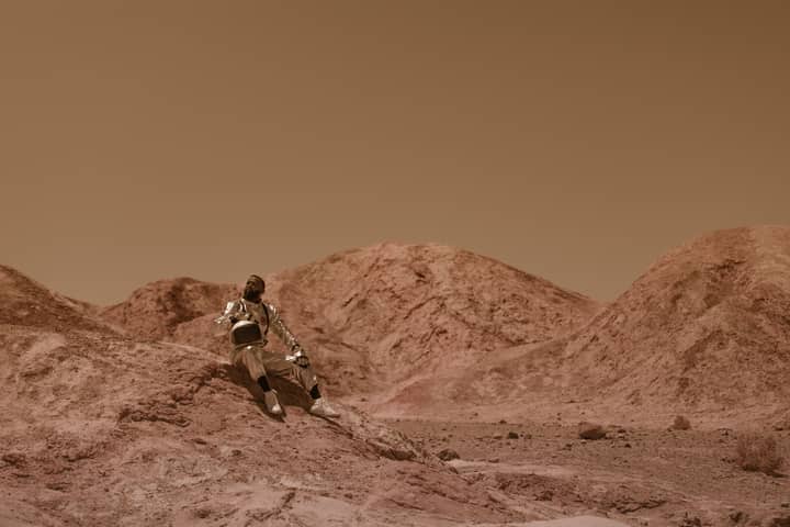 Astronaut in space suit sits on rocky desert terrain, evoking alien exploration.