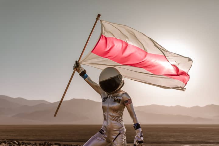 Astronaut in spacesuit holding a red and white flag in a desert setting with mountains in the background.