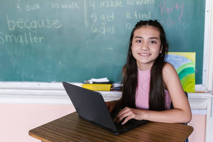 Young girl in a classroom, smiling while using a laptop, against a chalkboard backdrop.