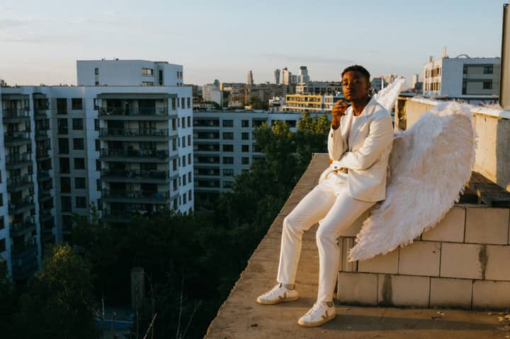 Man dressed as an angel with wings sitting on a rooftop at sunset, overlooking a cityscape.
