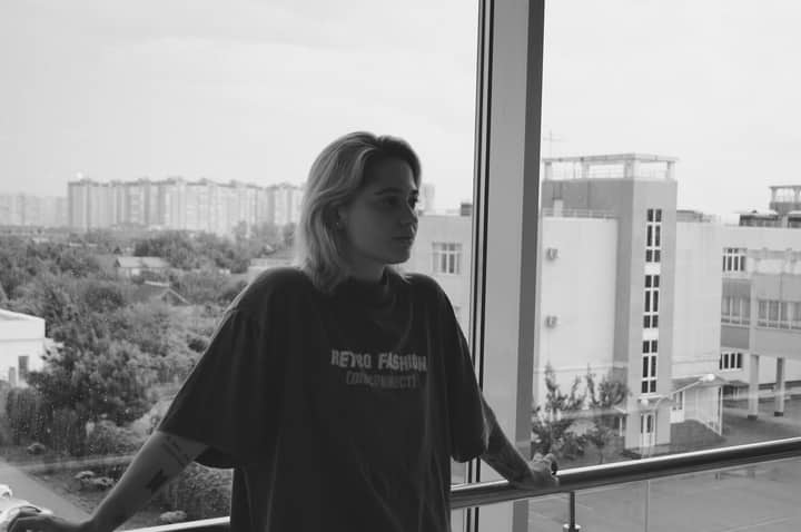 A young woman stands by a window, gazing out thoughtfully over a cityscape.