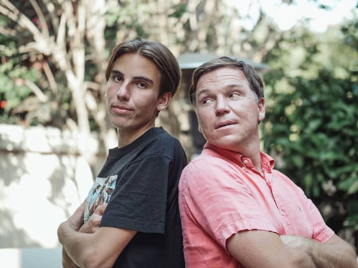 A father and son stand back to back, showcasing a playful bond in an outdoor setting.