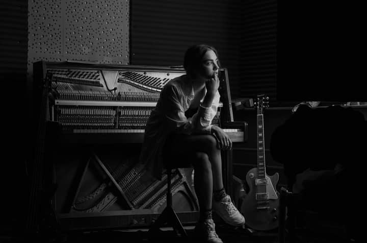 Black and white photo of a woman sitting by a piano in a studio, creating a dramatic and artistic atmosphere.