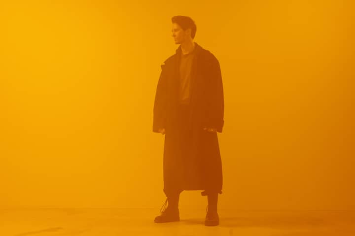 Man in black coat standing against an orange background, embodying a cyberpunk retro-futuristic style.