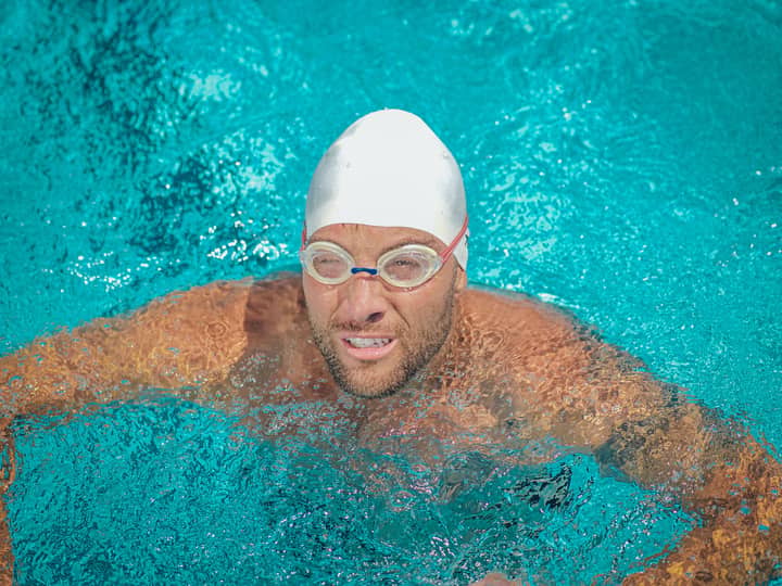 Close-up photo of a male swimmer with a white swimming cap and goggles in a clear blue pool.