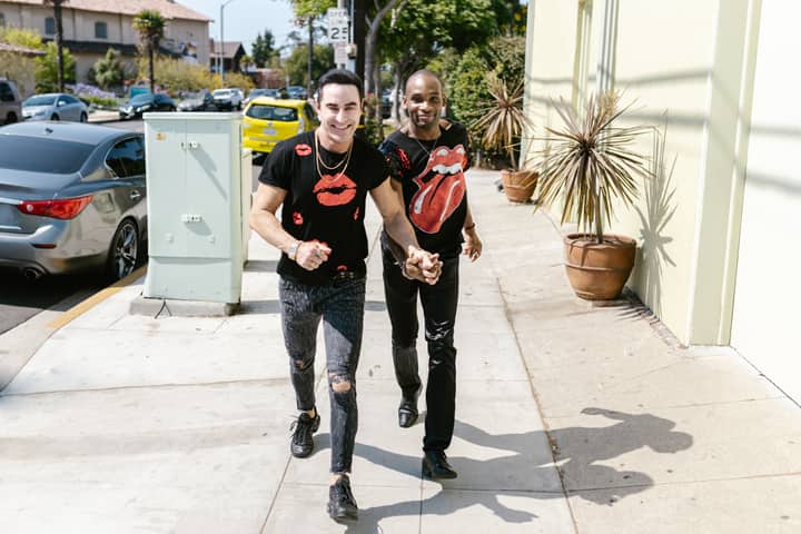 Smiling interracial gay couple holding hands while walking on a sunny urban sidewalk.