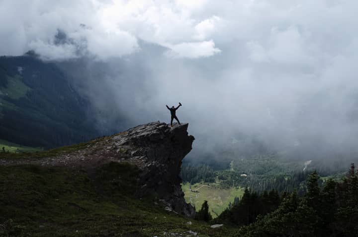 A lone hiker triumphantly stands on a misty mountain cliff, surrounded by clouds and forests.
