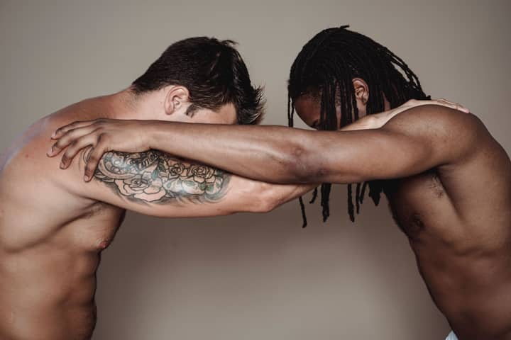 Two muscular men with intertwined arms in a studio setting, showing strength and diversity.