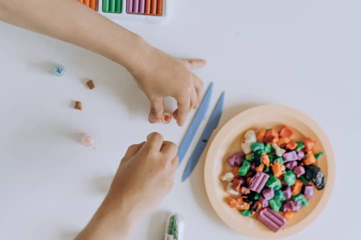 Hands crafting with colorful clay on a white surface, top view with artistic tools.