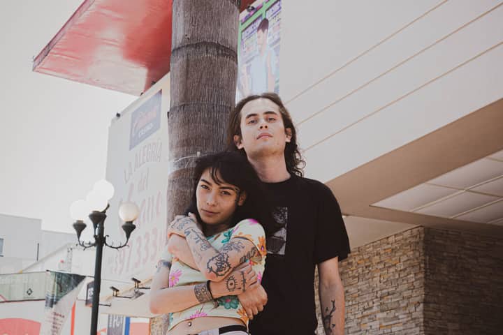 A stylish young couple embraces in an urban outdoor setting, showcasing tattoos and streetwear fashion.