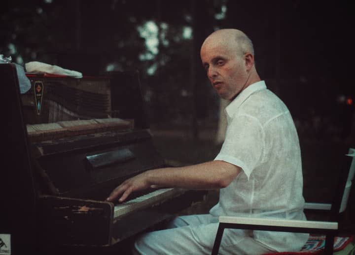 A bald man in a white shirt plays an old piano outdoors during twilight.