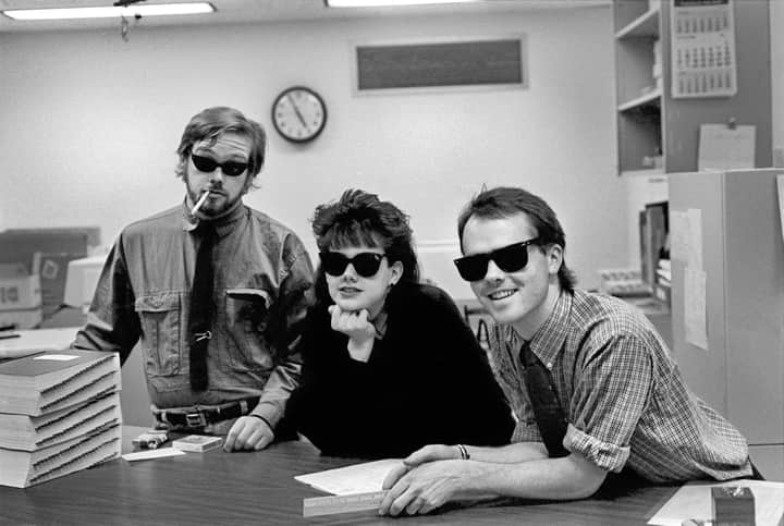 Black and white photo of three coworkers in an office wearing sunglasses, displaying a playful vibe.