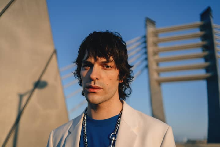 Stylish adult male wearing a beige blazer and blue shirt in front of modern bridge architecture.