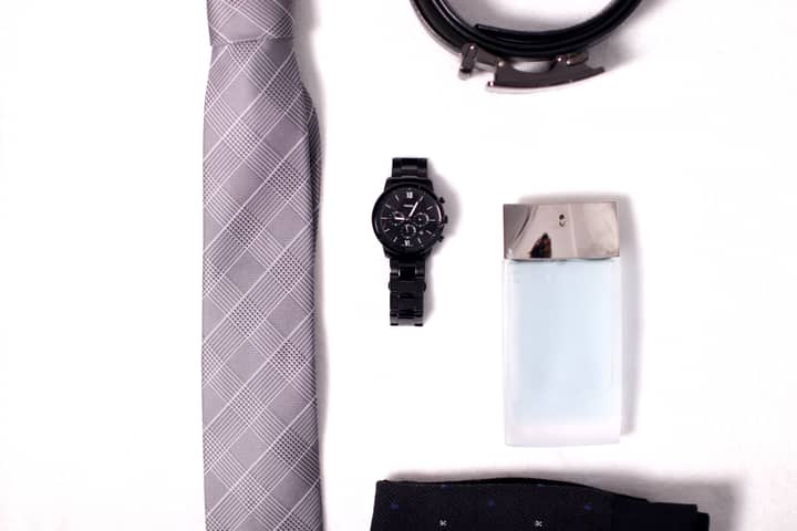 A flat lay of sophisticated men's accessories including a tie, watch, belt, and perfume on a white background.