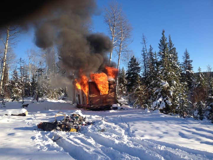 A cabin engulfed in flames amidst a snowy forest, depicting intense fire against the winter landscape.