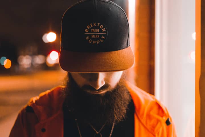 Bearded man in cap looking down, captured at night with urban lights in the background.