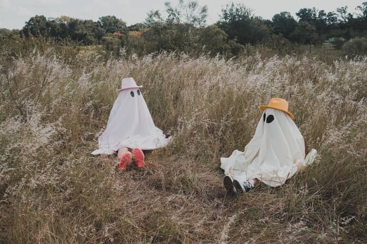 Two people dressed as ghosts with hats in a grassy field for Halloween fun.