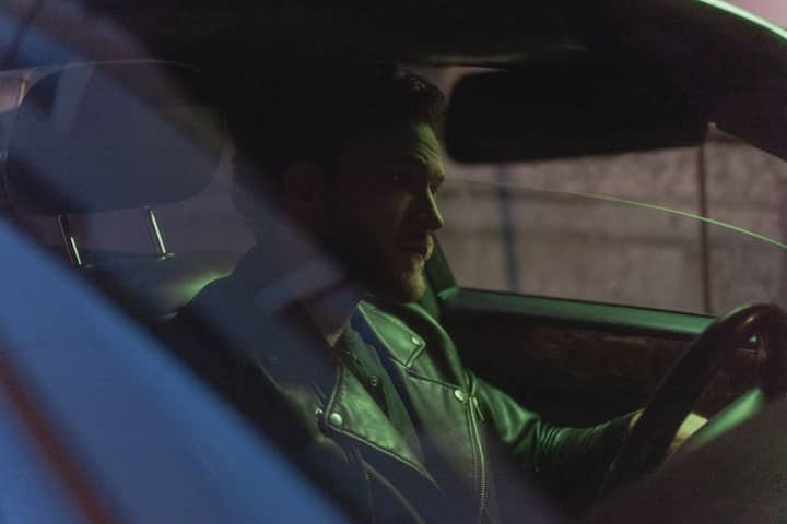 A bearded man in a leather jacket sits in a car, driving through the night.