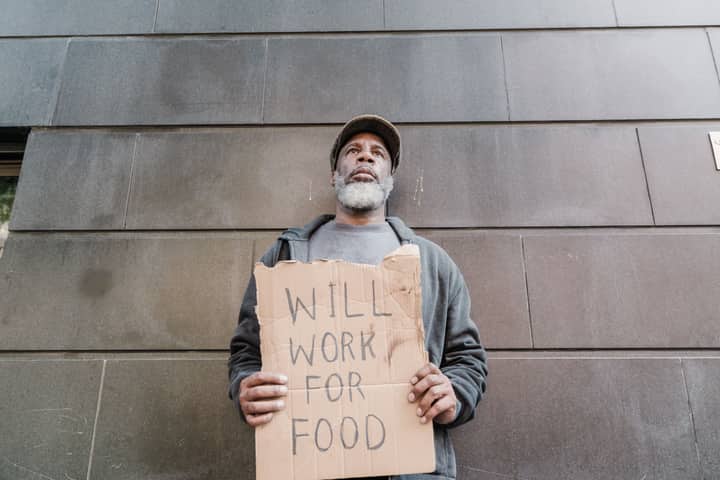 Bearded man on street holding a cardboard sign reading “Will Work for Food” against a gray wall.