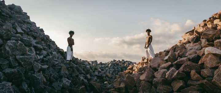 Two shirtless men stand among rocky hills under a dramatic sky.