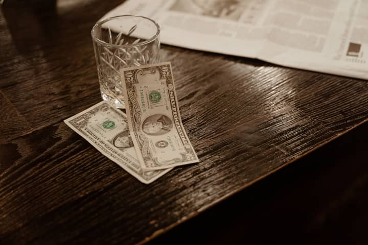 Close-up view of dollar bills with a glass on a wooden table, beside a newspaper.