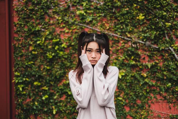Portrait of a young woman with pigtails in a sweater posing thoughtfully against a lush green ivy wall.