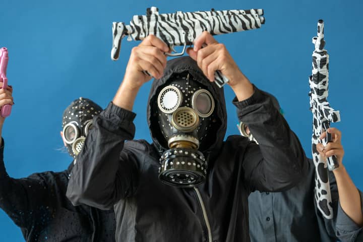 Three individuals in gas masks pose with toy guns against a blue backdrop.