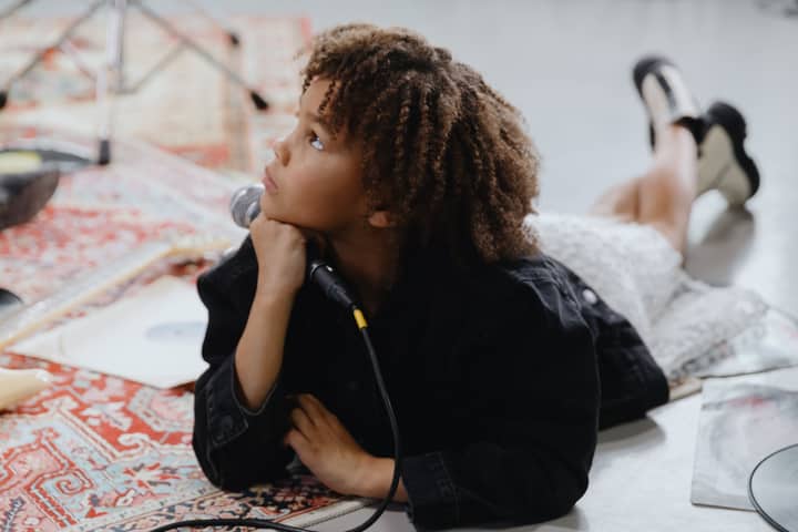 Curly-haired child lies indoors with a microphone, looking thoughtfully upwards.