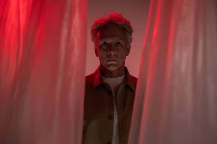 Elderly man with short gray hair looks through curtains under dramatic red lighting. Studio portrait.