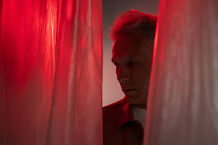 Elderly man in dark, dramatic lighting with red curtains, gazing thoughtfully.