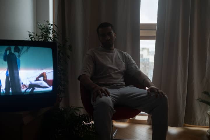 A man sits indoors watching TV, curtains drawn, creating a moody atmosphere.