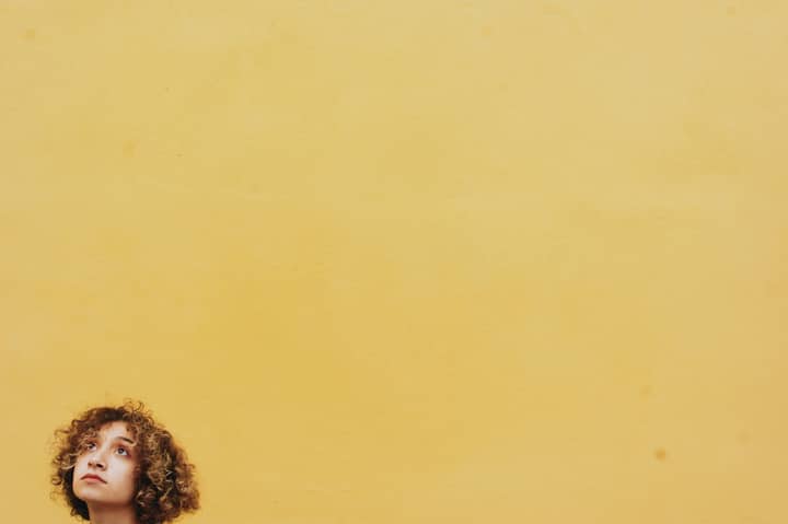 Portrait of a curly-haired woman looking up against a vibrant yellow background with space for text.