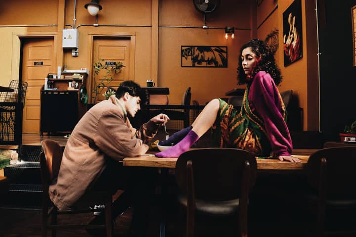 A fashionable couple sitting in a trendy cafe with colorful decor, showcasing unique poses and attire.