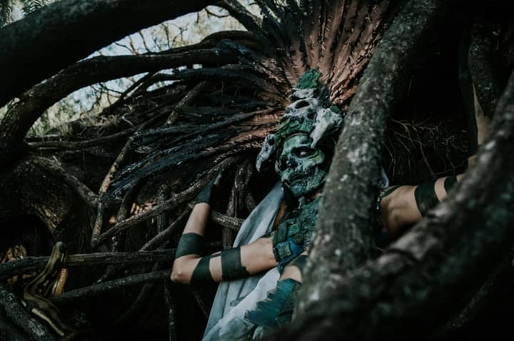 A person in an elaborate headdress and mask blends with twisted tree branches, evoking mythological themes.